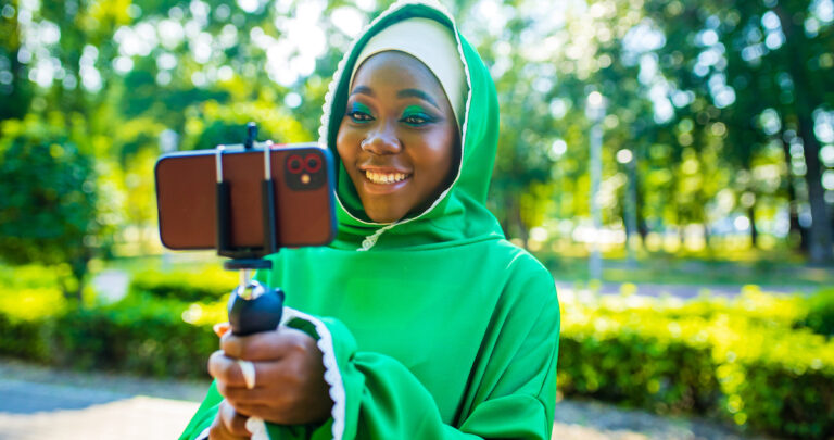 latin hispanic wonan in green muslim hijab with bright make up and nose piercing taking video stream outdoors summer park