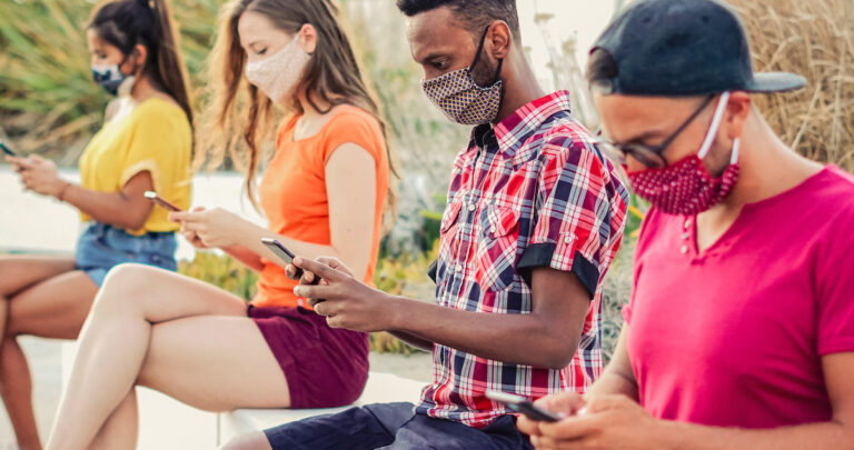 Friends using their smartphones in covid 19 times protected with face mask - Young people using mobile device in distance outdoors